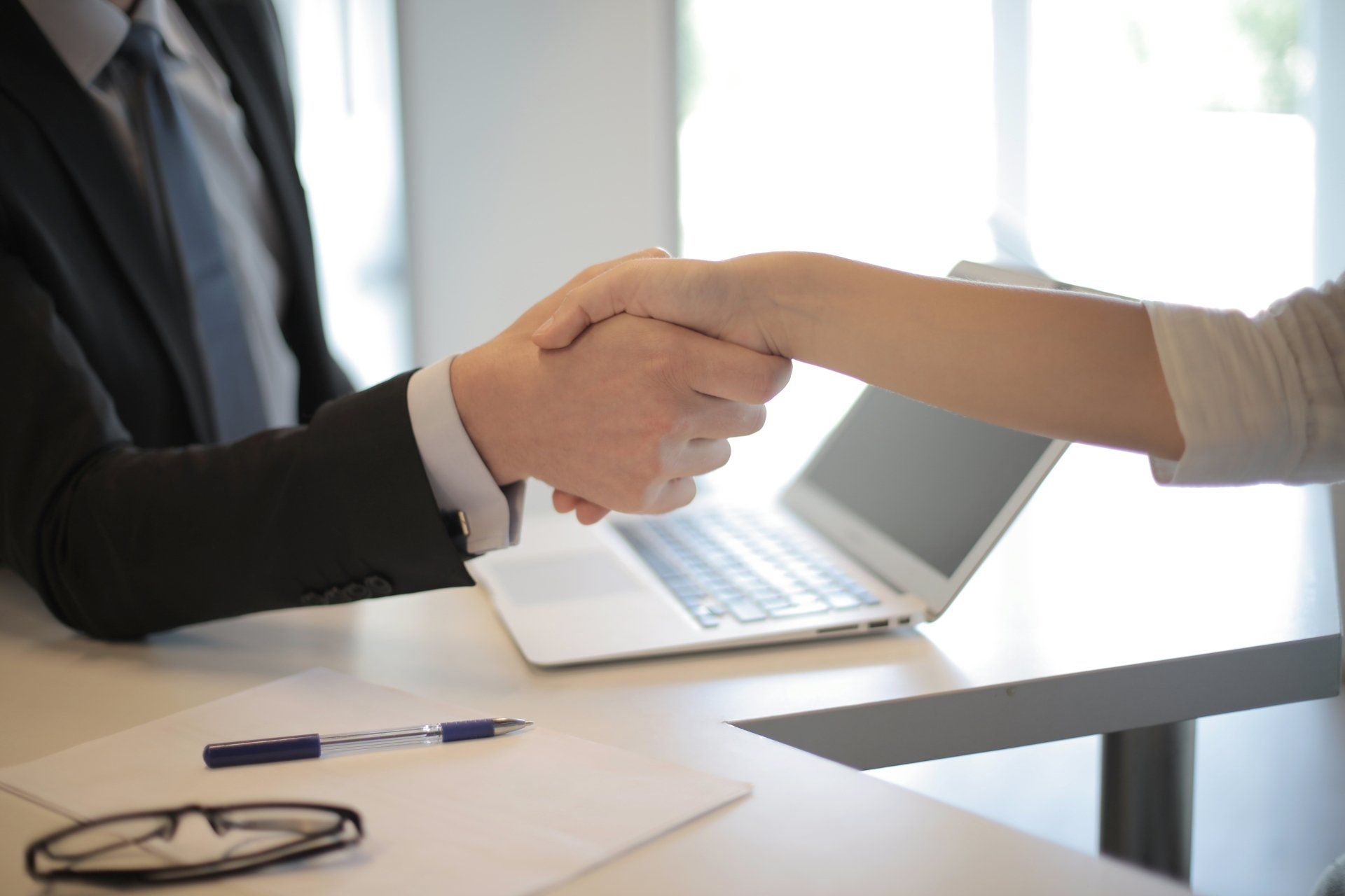 Two people shaking hands over a laptop at a desk in a bright office